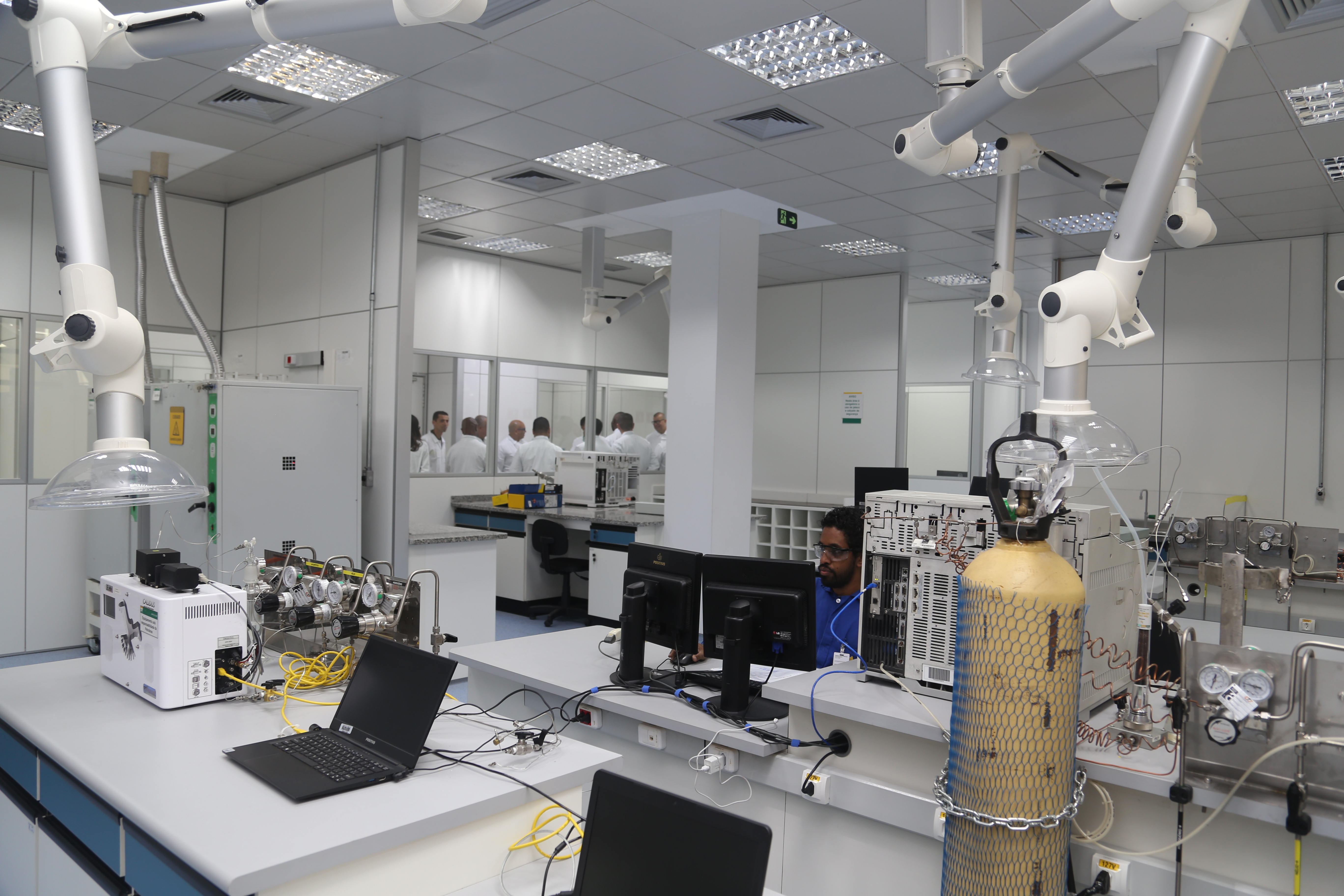 Sala do laboratório de fluidos, com equipamentos, notebooks e monitores sobre bancadas e um pesquisador de camisa azul, sentado ao fundo, olhando para um dos monitores